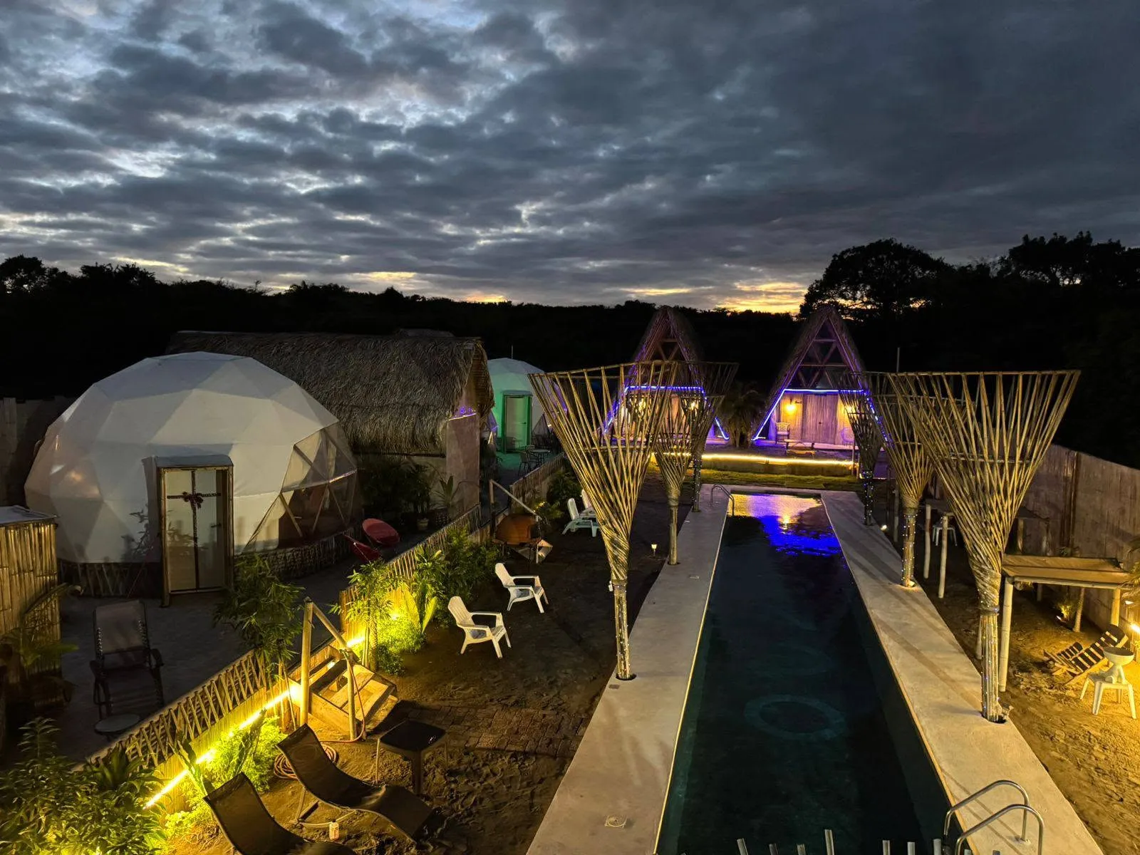 Vista aérea del glamping al atardecer: domos, cabañas tipo A y la alberca junto a las dunas