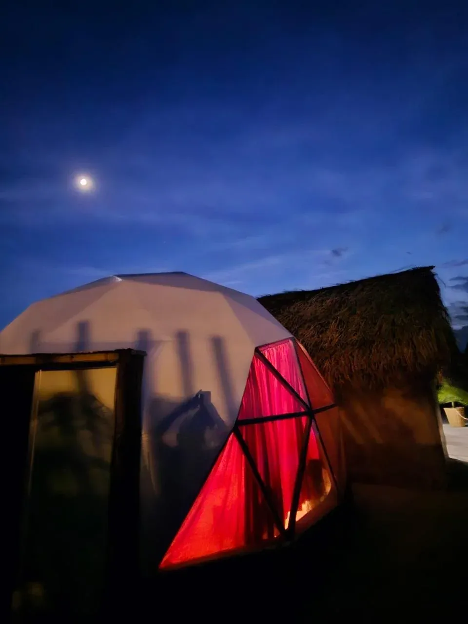 Domo geodésico con ventanas rojas bajo la luna, al anochecer