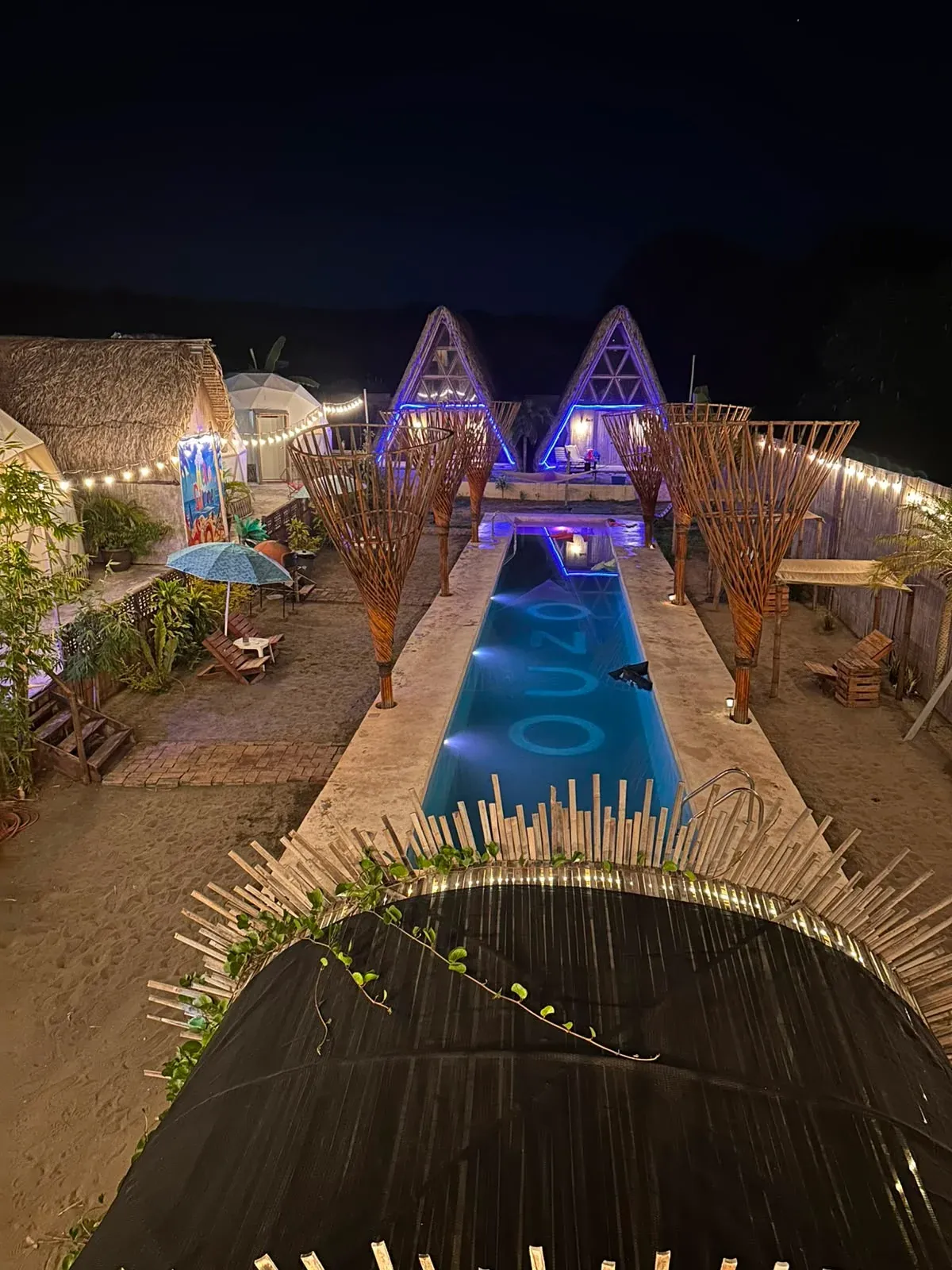 El glamping de noche: la alberca iluminada entre esculturas de bambú, con las cabañas tipo A y un domo al fondo